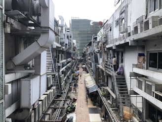 A narrow alleyway flanked by buildings with multiple air conditioning units and metal ducts attached to the exterior walls. The alley is cluttered with outdoor staircases, parked motorcycles, and hanging laundry. There is a mixture of concrete and corrugated metal structures, with a slight view of a larger glass building in the background.