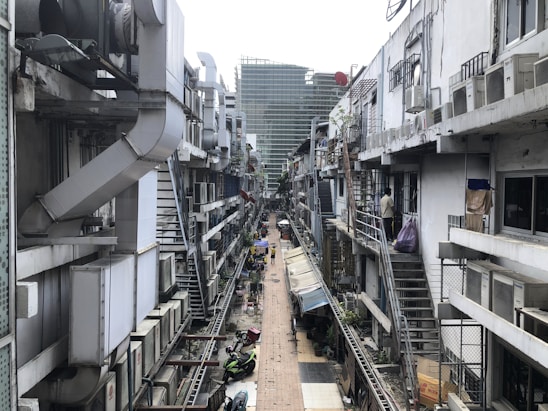 A narrow alleyway flanked by buildings with multiple air conditioning units and metal ducts attached to the exterior walls. The alley is cluttered with outdoor staircases, parked motorcycles, and hanging laundry. There is a mixture of concrete and corrugated metal structures, with a slight view of a larger glass building in the background.