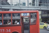 A red public bus is moving through a city street. The bus has number 77 displayed, and passengers are seated inside, some using their phones. The windows of the bus have text in a foreign language. In the background, there is a pedestrian bridge and another vehicle, a yellow taxi.