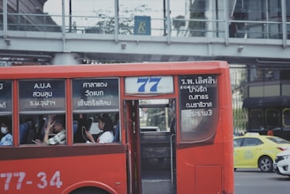 A red public bus is moving through a city street. The bus has number 77 displayed, and passengers are seated inside, some using their phones. The windows of the bus have text in a foreign language. In the background, there is a pedestrian bridge and another vehicle, a yellow taxi.