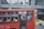 A red public bus is moving through a city street. The bus has number 77 displayed, and passengers are seated inside, some using their phones. The windows of the bus have text in a foreign language. In the background, there is a pedestrian bridge and another vehicle, a yellow taxi.