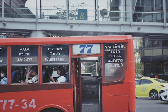 A red public bus is moving through a city street. The bus has number 77 displayed, and passengers are seated inside, some using their phones. The windows of the bus have text in a foreign language. In the background, there is a pedestrian bridge and another vehicle, a yellow taxi.