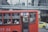 A red public bus is moving through a city street. The bus has number 77 displayed, and passengers are seated inside, some using their phones. The windows of the bus have text in a foreign language. In the background, there is a pedestrian bridge and another vehicle, a yellow taxi.