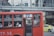 A red public bus is moving through a city street. The bus has number 77 displayed, and passengers are seated inside, some using their phones. The windows of the bus have text in a foreign language. In the background, there is a pedestrian bridge and another vehicle, a yellow taxi.