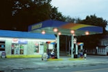 A small gas station with a convenience store, featuring a blue and yellow canopy displaying the Sunoco logo. Two gas pumps are visible under the brightly lit canopy. The store windows are filled with signs and advertisements, and the surrounding area includes trees and a residential building.