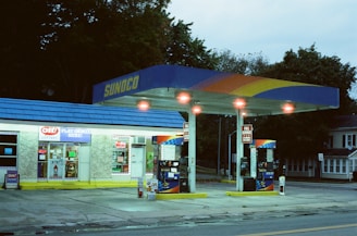 A small gas station with a convenience store, featuring a blue and yellow canopy displaying the Sunoco logo. Two gas pumps are visible under the brightly lit canopy. The store windows are filled with signs and advertisements, and the surrounding area includes trees and a residential building.
