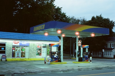 A small gas station with a convenience store, featuring a blue and yellow canopy displaying the Sunoco logo. Two gas pumps are visible under the brightly lit canopy. The store windows are filled with signs and advertisements, and the surrounding area includes trees and a residential building.