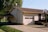 A suburban house with a sloped roof features a garage with two windows. A basketball hoop is mounted on a pole near the garage entrance. The house siding is light-colored, with a small set of steps leading to a dark-colored front door. Adjacent to the house is a carport and a driveway, with green lawns and trees surrounding the property. Cables and a satellite dish are visible on the side of the house.
