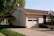 A suburban house with a sloped roof features a garage with two windows. A basketball hoop is mounted on a pole near the garage entrance. The house siding is light-colored, with a small set of steps leading to a dark-colored front door. Adjacent to the house is a carport and a driveway, with green lawns and trees surrounding the property. Cables and a satellite dish are visible on the side of the house.