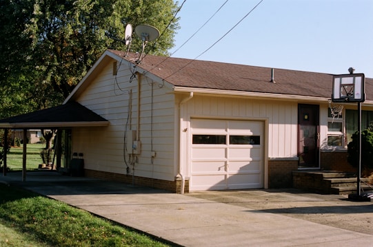 A suburban house with a sloped roof features a garage with two windows. A basketball hoop is mounted on a pole near the garage entrance. The house siding is light-colored, with a small set of steps leading to a dark-colored front door. Adjacent to the house is a carport and a driveway, with green lawns and trees surrounding the property. Cables and a satellite dish are visible on the side of the house.