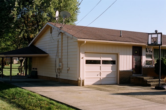 A suburban house with a sloped roof features a garage with two windows. A basketball hoop is mounted on a pole near the garage entrance. The house siding is light-colored, with a small set of steps leading to a dark-colored front door. Adjacent to the house is a carport and a driveway, with green lawns and trees surrounding the property. Cables and a satellite dish are visible on the side of the house.