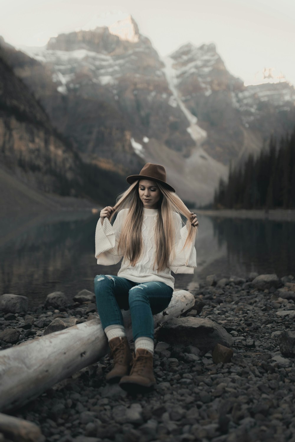 woman in white jacket and blue denim jeans sitting on rock near lake during daytime