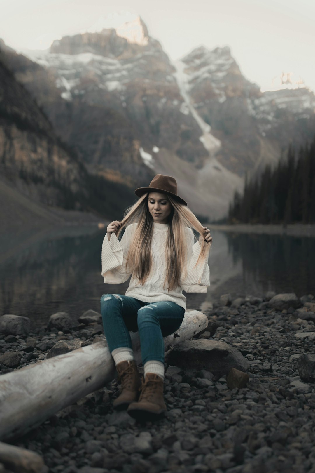 woman in white jacket and blue denim jeans sitting on rock near lake ...