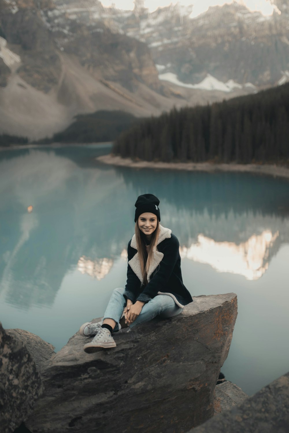 Frau in schwarzer Strickjacke sitzt tagsüber auf Felsen in der Nähe des Sees
