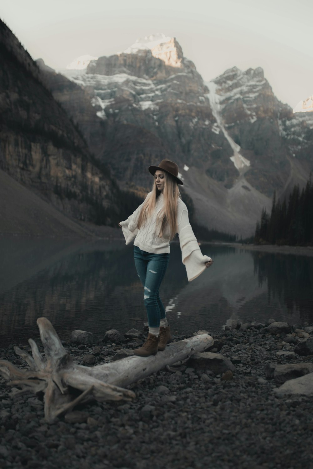 woman in white jacket and blue denim jeans standing on rock near lake during daytime