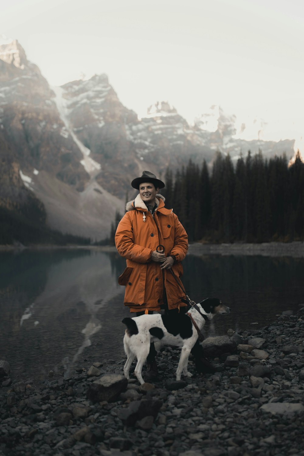 man in brown jacket standing beside dog near lake during daytime