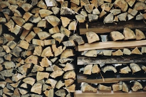 A neatly stacked pile of chopped firewood, with logs showing various shades of brown and gray. The logs are arranged tightly together, forming a rustic and natural pattern.