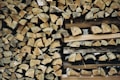 A neatly stacked pile of chopped firewood, with logs showing various shades of brown and gray. The logs are arranged tightly together, forming a rustic and natural pattern.