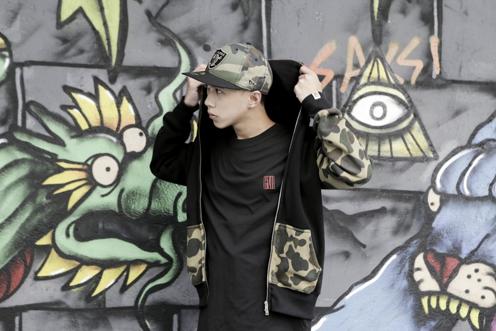 man in black and white adidas hoodie standing beside wall with graffiti