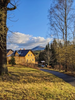 A charming country house with a yellow facade and a dark roof is set against a backdrop of tall, leafless trees and distant mountains. The sky is clear and bright blue with scattered fluffy clouds. A narrow paved path meanders alongside a grassy field, bathed in warm, golden sunlight.