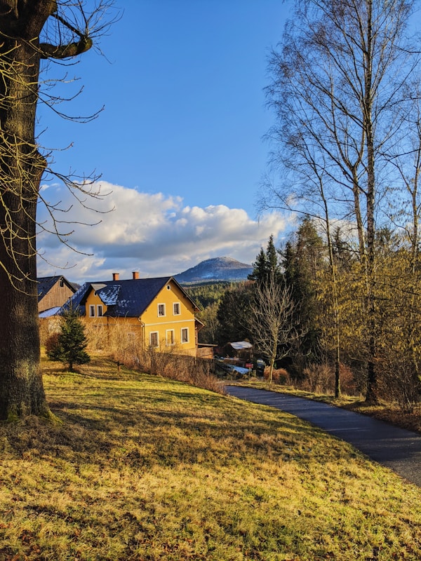 A charming country house with a yellow facade and a dark roof is set against a backdrop of tall, leafless trees and distant mountains. The sky is clear and bright blue with scattered fluffy clouds. A narrow paved path meanders alongside a grassy field, bathed in warm, golden sunlight.