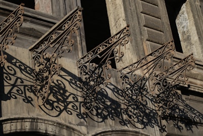 Intricate wrought iron railings cast complex, swirling shadows on the façade of an old stone building. The architecture features detailed craftsmanship with ornamental designs that extend from the balcony.