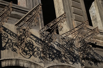 Intricate wrought iron railings cast complex, swirling shadows on the façade of an old stone building. The architecture features detailed craftsmanship with ornamental designs that extend from the balcony.