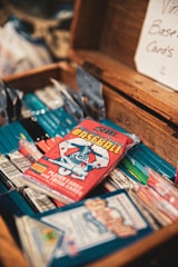 A collection of vintage baseball card packs is arranged inside an open wooden box. The cards are colorful, with a focus on a red pack featuring an illustration of a baseball player. A sign in the background reads 'Vintage Baseball Cards'.