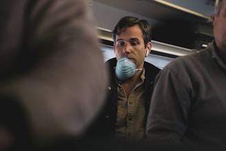 A man wearing a face mask and headphones is seated in an aircraft cabin. He appears contemplative or perhaps concerned, surrounded by other passengers whose faces are partially visible. The lighting is dim, suggesting an indoor setting during a flight.