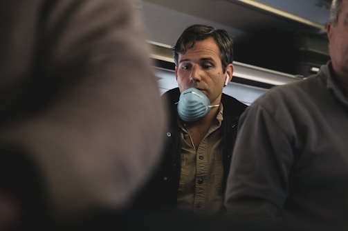 A man wearing a face mask and headphones is seated in an aircraft cabin. He appears contemplative or perhaps concerned, surrounded by other passengers whose faces are partially visible. The lighting is dim, suggesting an indoor setting during a flight.