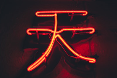 A bright red neon sign displaying a complex symbol against a dark background. The glowing neon tubing is intricately shaped and emits a vivid light, creating a stark contrast with the dim surroundings.