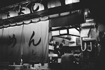 A black and white photograph of a traditional Japanese food stall with hanging lanterns above and large cloth banners with Japanese characters. Inside the stall, there are two people working, one partially visible in the background. Various kitchen equipment and supplies are seen in the workspace.