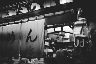 A black and white photograph of a traditional Japanese food stall with hanging lanterns above and large cloth banners with Japanese characters. Inside the stall, there are two people working, one partially visible in the background. Various kitchen equipment and supplies are seen in the workspace.