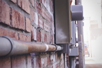A close-up view of a weathered brick wall with a metal conduit attached. The perspective shows the conduit running parallel to the wall. Metal electrical boxes and other utility equipment are mounted beside the brick wall. The image captures an industrial and urban setting with utility poles and blurred background indicating an alleyway.