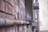 A close-up view of a weathered brick wall with a metal conduit attached. The perspective shows the conduit running parallel to the wall. Metal electrical boxes and other utility equipment are mounted beside the brick wall. The image captures an industrial and urban setting with utility poles and blurred background indicating an alleyway.