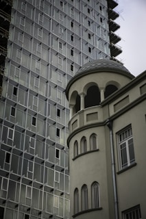 A modern high-rise building with a grid-like pattern of windows sits next to a more classic structure featuring arched windows and a rounded tower. The contrast in architectural styles is notable, highlighting both contemporary and traditional elements.
