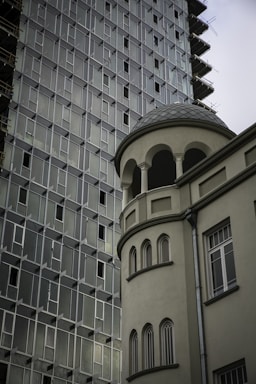 A modern high-rise building with a grid-like pattern of windows sits next to a more classic structure featuring arched windows and a rounded tower. The contrast in architectural styles is notable, highlighting both contemporary and traditional elements.
