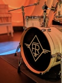 A close-up view of a drum set focusing on a large bass drum with a distinctive logo designed with stylized letters 'FR' inside a diamond shape. The drum is set up on a stage with muted lighting, producing a warm ambiance.