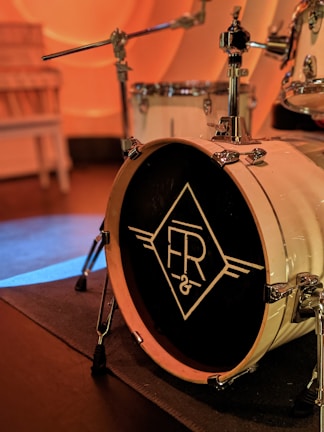 A close-up view of a drum set focusing on a large bass drum with a distinctive logo designed with stylized letters 'FR' inside a diamond shape. The drum is set up on a stage with muted lighting, producing a warm ambiance.
