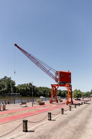 A large red industrial crane is positioned on a riverside promenade, surrounded by neatly arranged pedestrian paths and bordered by chains. The area features several benches, flower planters, and a distant boat visible on the water. Trees and greenery line the opposite bank under a clear blue sky.