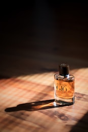 A bottle of Acqua di Giò Giorgio Armani perfume is placed on a wooden surface with sunlight streaming through, casting shadows around the bottle. The liquid inside the clear glass bottle is amber in color, and the bottle has a dark cap.