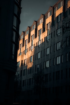 A multi-story building with a series of windows is partially illuminated by the warm glow of sunlight. The light creates a striking contrast against the shadowed portions of the building, highlighting its modern architectural design. The angular rooftops and reflections in the glass give the structure a dynamic appearance.