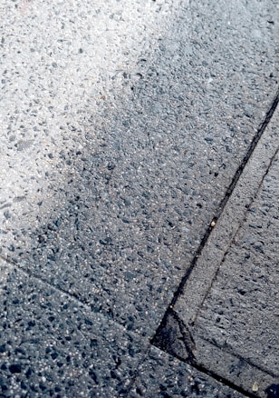 The image displays a close-up view of wet asphalt with visible texture. The surface exhibits a mix of light and dark tones, with small pebbles embedded throughout. There are faint outlines of rectangular shapes, possibly indicating pavement sections.