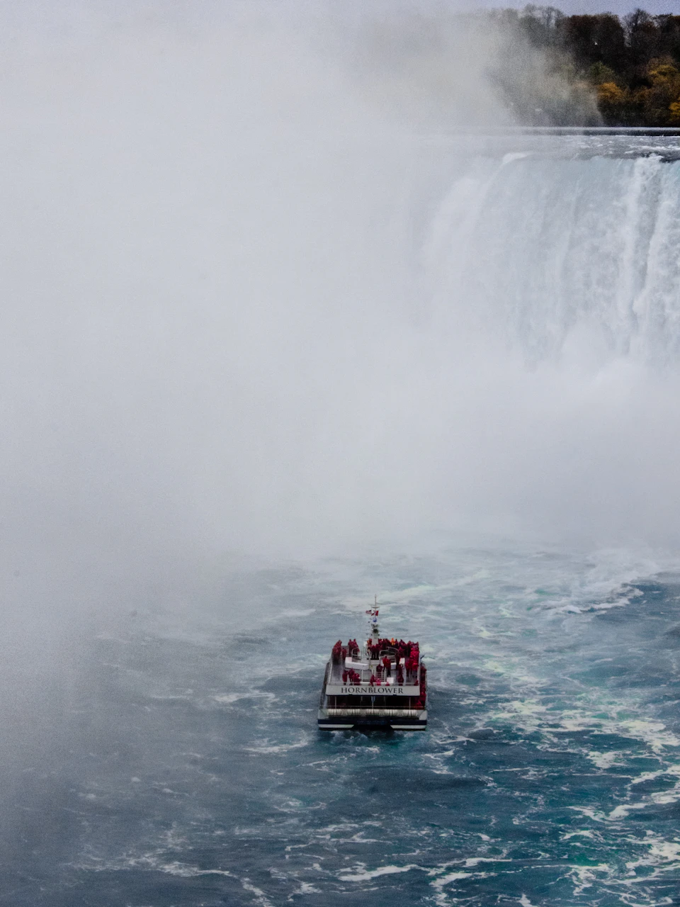 The image depicts a boat floating on a river, with a group of people aboard it. The boat is painted red and white and has a mast extending from its side. The river flows through a mountainous landscape, creating a misty atmosphere around the scene. In the distance, a waterfall can be seen, adding to the natural beauty of the location.