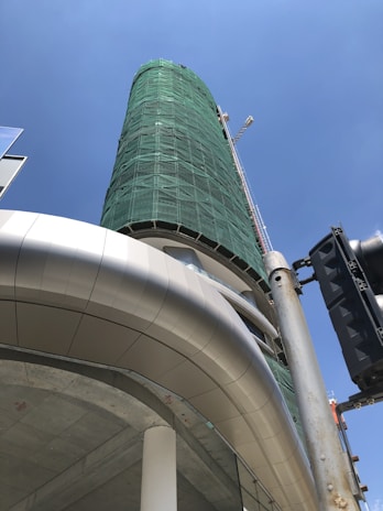 A tall cylindrical building under construction is wrapped in green netting for safety. The view is from a low angle, showcasing the curvature of the structure. A construction crane is visible alongside the building, as well as a traffic signal in the foreground. Part of the structure includes a modern, metallic architectural element.