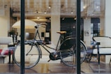 A vintage-style bicycle is placed inside a modern showroom with large windows. The interior features comfortable, minimalist furniture including chairs and a sofa. Soft lighting creates a cozy and inviting atmosphere with an elegant, urban aesthetic.