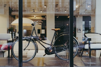 A vintage-style bicycle is placed inside a modern showroom with large windows. The interior features comfortable, minimalist furniture including chairs and a sofa. Soft lighting creates a cozy and inviting atmosphere with an elegant, urban aesthetic.