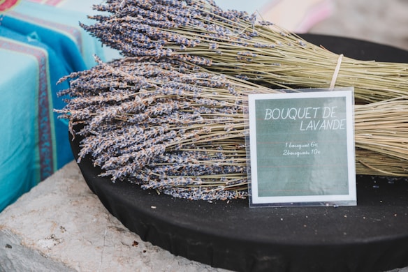 Bundles of dried lavender are arranged on a dark surface accompanied by a sign indicating prices for single and multiple bouquets. The background features a stone surface and a turquoise cloth.