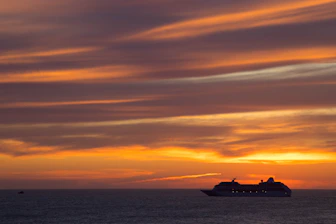 A luxurious cruise ship sailing at sunset with vibrant orange and pink skies.