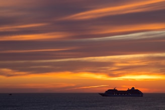 A large cruise ship travels on the ocean against a backdrop of a vibrant sunset. The sky is filled with streaks of orange, yellow, and purple clouds, creating a dramatic and serene atmosphere.